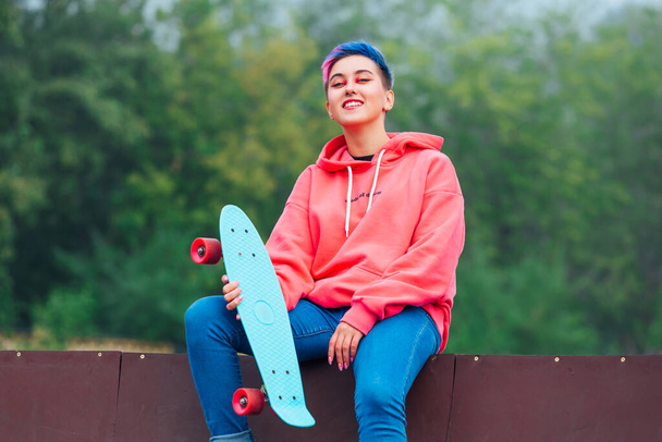 Summer lifestyle image of trendy pretty young girl with short colored hair and nose piercing dressed in pink hoodie and jeans sitting next to the skateboard coart with her blue plastic skateboard. - Foto, Imagem