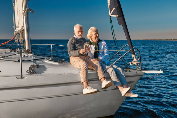 Celebrando el aniversario de boda. Feliz pareja de ancianos bebiendo vino o champán y riéndose mientras están sentados en un velero o yate flotando en un tranquilo mar azul - Foto, Imagen