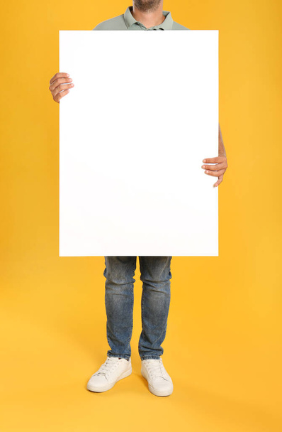 Мужчина держит белый пустой плакат на желтом фоне, крупным планом. Макет для дизайна - Фото, изображение