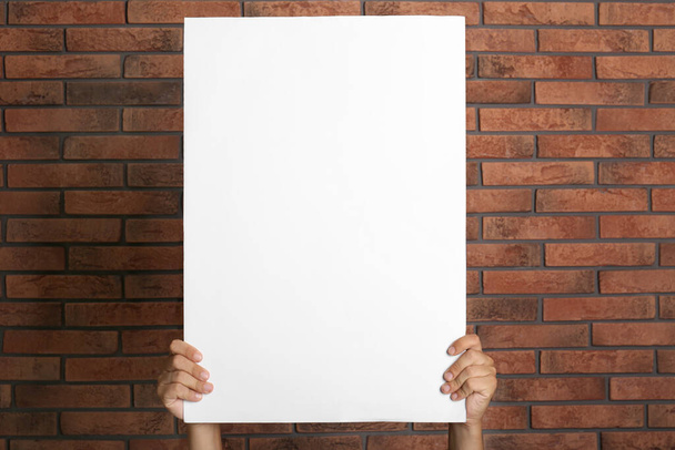 Man holding white blank poster near red brick wall, closeup. Mockup for design - Fotó, kép
