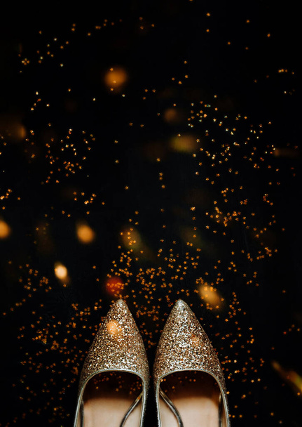 goud glanzende vrouwen vakantie puntige schoenen op zwarte achtergrond en glanzende confetti sterren rond, vakantie achtergrond, selectieve focus, verticale - Foto, afbeelding