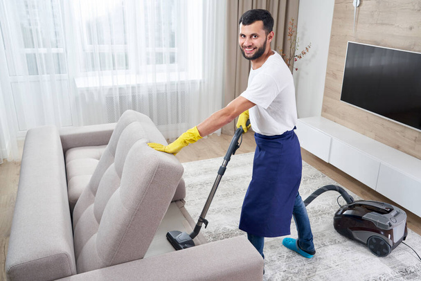 Giovane uomo Divano di pulizia con aspirapolvere in lasciare la stanza a casa - Foto, immagini