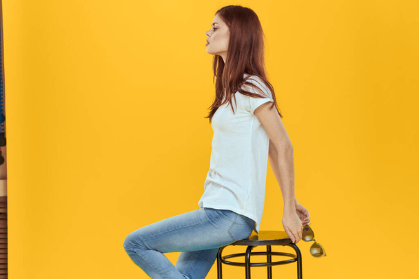 椅子に座っている女性白いTシャツのジーンズファッショナブルな服魅力的なスタジオ黄色の背景 - 写真・画像