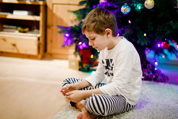 Cute little blond kid boy using smartphone on Christmas with decorated tree on background. Happy healthy hild having fun at home. Popular gift for xmas - Foto, imagen