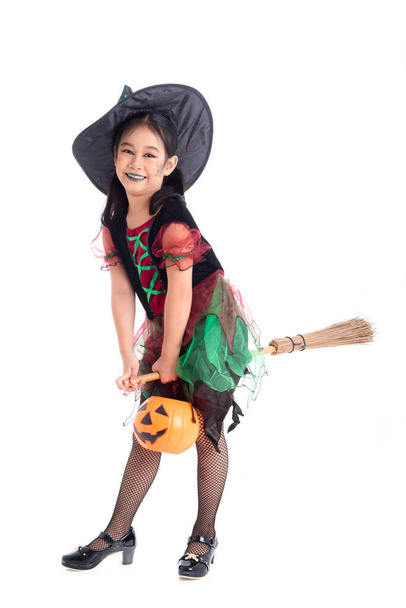 Volledige lengte van kleine Aziatische meisje in heks kostuum met make-up rijden op bezem en glimlacht over witte achtergrond. Kind Halloween kostuum concept. - Foto, afbeelding