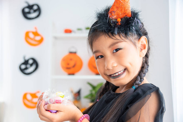 Little asian girl in witch dress smiling and holding many candy on hands in halloween decorated room. - Фото, изображение