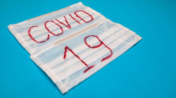 Typical white 3-ply surgical mask with rubber earhooks to cover mouth and nose with English block letters Covid-19 on a blue background. Dangerous virus and quarantine concept - Photo, Image