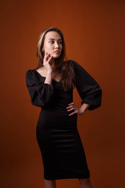 элегантная модель в черном платье с глубоким вырезом позирует на оранжевом фоне - Фото, изображение