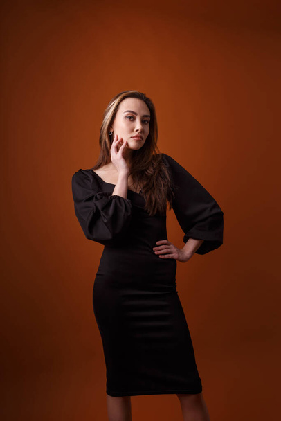 элегантная модель в черном платье с глубоким вырезом позирует на оранжевом фоне - Фото, изображение
