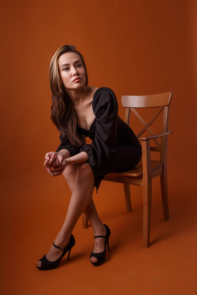 grazioso modello in abito nero, scarpe tacco alto siede in sedia su sfondo arancione - Foto, immagini
