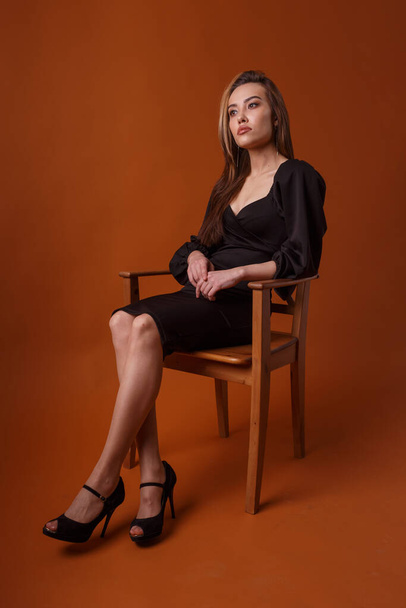 hübsches Model im schwarzen Kleid, hohe Schuhe sitzt im Stuhl auf orangefarbenem Hintergrund - Foto, Bild