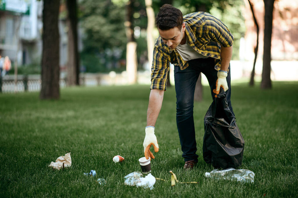 Young man collects garbage in a bag in park, volunteering. Male person cleans forest, ecological restoration, eco lifestyle, trash collection and recycling, ecology care, cleaning - Photo, Image