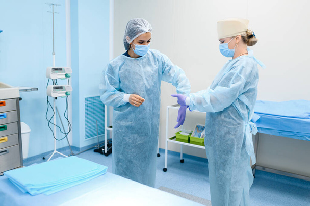Chirurgin und Assistentin ziehen im Operationssaal Handschuhe an und bereiten sich auf die Operation vor. Arzt in Uniform, medizinisches Klinikpersonal, Medizin und Gesundheit, Gesundheitsfürsorge im Krankenhaus - Foto, Bild