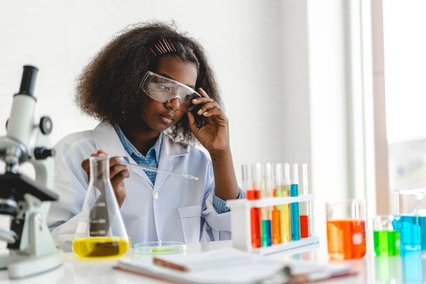Afrika kökenli sevimli küçük kız öğrenci araştırma yapmayı ve kimyasal deney yapmayı öğrenirken fen bilimleri sınıfında sıvıyı analiz edip karıştırıyor. Eğitim ve bilim konsepti. - Fotoğraf, Görsel
