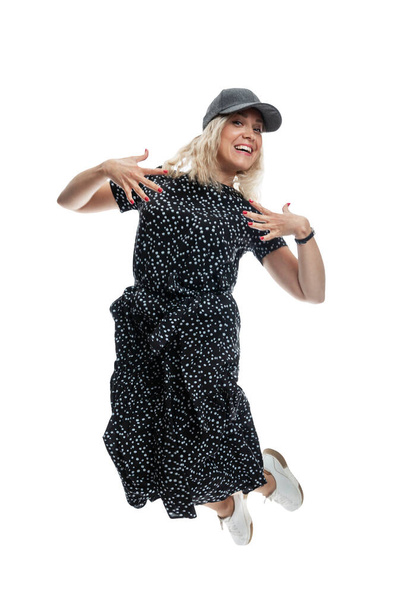 A laughing young girl in a black dress, a cap and white sneakers is jumping. Freedom and relaxation. Isolated on white background. Vertical. - Fotoğraf, Görsel
