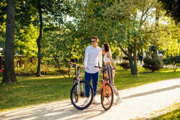 Ritratto a figura intera di un bellissimo giovane uomo e donna in piedi nel parco con le biciclette e distogliendo lo sguardo con un sorriso sul viso. Elegante coppia con biciclette trascorre un tempo attivo nel parco. - Foto, immagini