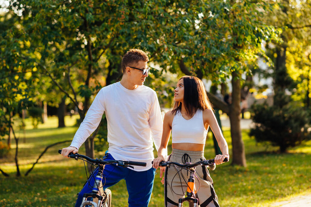 Stylish couple in love on bicycles in the autumn park looking at each other with a serious face. Portrait of stylish woman and man on bicycles on a walk outdoors. - Foto, Imagem