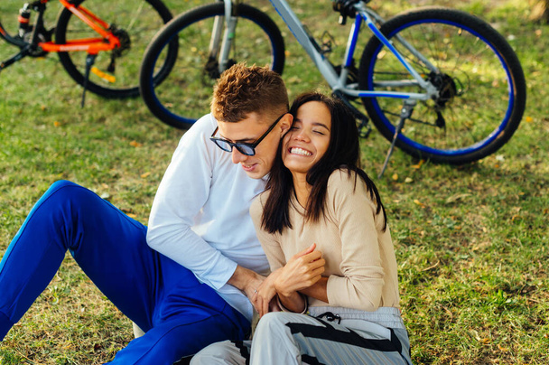 公園の芝生の上に座って楽しみを持っている愛の幸せなカップル、男は女性をくすぐる、彼女は笑顔。自転車を背景に芝生の上でスタイリッシュな若いカップルのレジャー. - 写真・画像