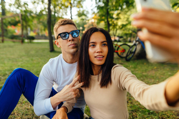 Krásný mladý pár sedí na trávníku v parku a bere selfie na smartphonu. Žena fotografování s přítelem při relaxaci v parku - Fotografie, Obrázek