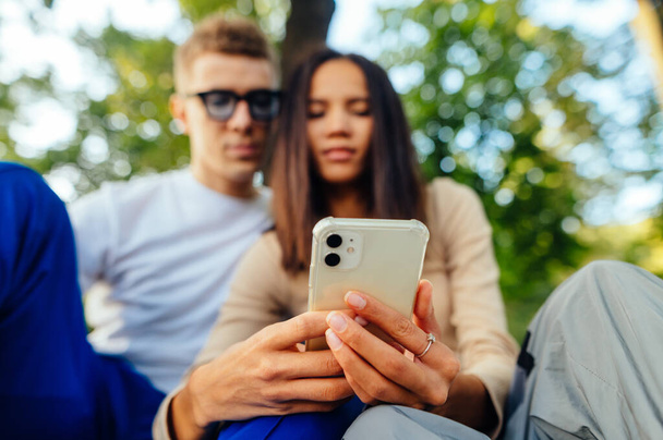 公園に座っているカップルやスマートフォンでインターネットを使用して、画面を見てください。手に携帯電話を持つ女性の手に焦点を当てる. - 写真・画像
