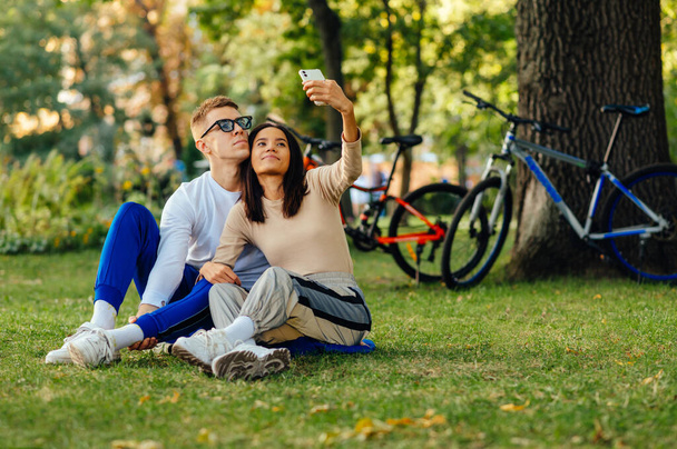 Beautiful young couple taking a selfie sitting in the park on the grass on a background of bicycles.Woman and man spend time together in the park, take a joint photo without a smartphone selfie camera - Photo, Image