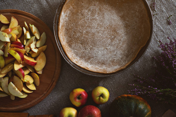 Preparation, Thanksgiving Day celebration. Concept. Top view of a pan with pie dough, a board with a slide of apples and pumpkins cut for pie on the gray linen tablecloth. - 写真・画像