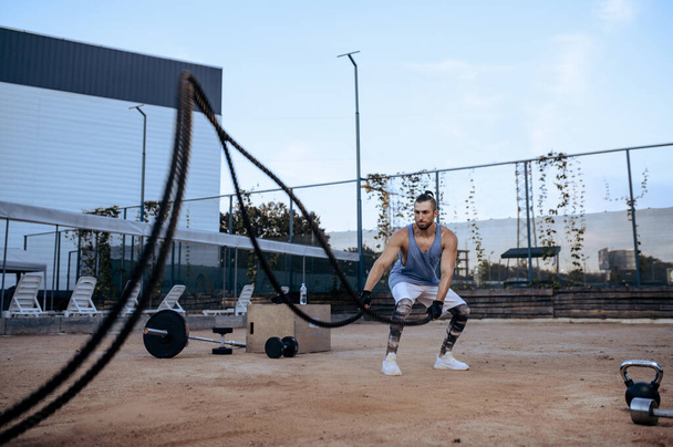Αθλητικός άνθρωπος κάνει άσκηση με σχοινιά, προπόνηση δρόμου, crossfit. Άσκηση γυμναστικής σε γήπεδα υπαίθρια, αρσενικό άτομο αντλεί μυς, ενεργό αστικό τρόπο ζωής - Φωτογραφία, εικόνα