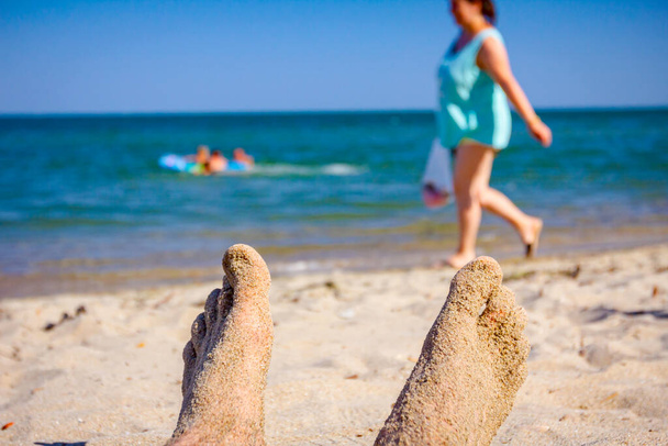Skrzyżowane nogi mężczyzny opalają się, leżąc beztrosko na piasku obok wybrzeża, na publicznej plaży.. - Zdjęcie, obraz