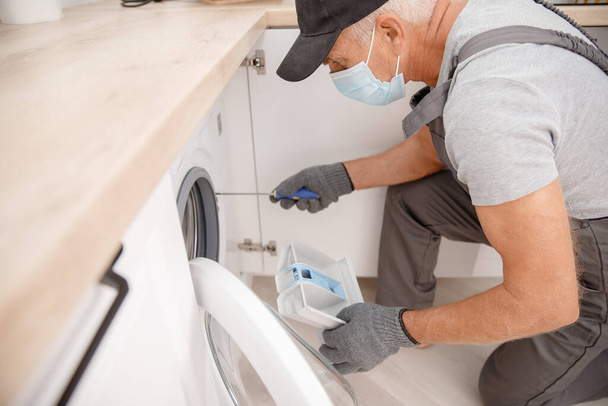 Orvosi maszkos vízvezeték szerelő javítgatja a mosógépet a konyhában. Koncepciószerviztechnikus - Fotó, kép