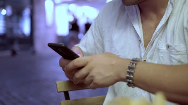 Zbliżenie rąk mężczyzn trzymających telefon komórkowy i wpisujących wiadomości na oświetlonym zatłoczonym tle miasta, mężczyzna siedzący na zewnątrz w kawiarni i korzystający ze smartfona, przeglądający internet lub komunikujący się z - Materiał filmowy, wideo