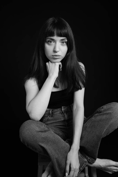 Studio portrait of a pretty barefoot brunette woman in black spaghetti strap top and jeans, looking at the camera, sitting, legs crossed, against a plain black background - Foto, imagen