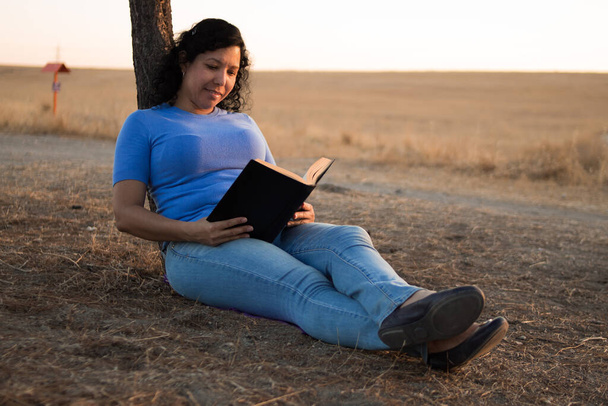 Fiatal kaukázusi nő olvas egy fekete könyvet ül a földön egy parkban naplementével a háttérben - Fotó, kép