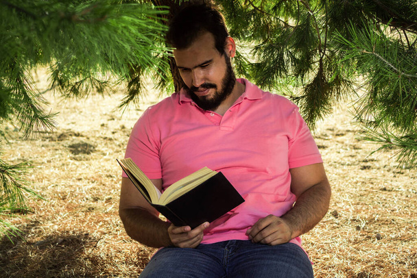 Hombre caucásico en pantalones vaqueros y una camisa rosa sentado cerca de un árbol en un bosque leyendo un libro negro - Foto, Imagen
