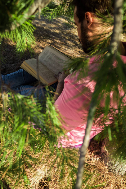 Uomo caucasico in jeans pantaloni e una camicia rosa seduto vicino a un albero in una foresta a leggere un libro nero - Foto, immagini