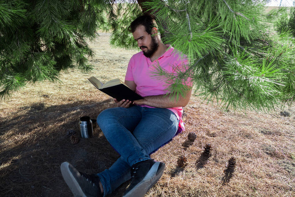 Hombre caucásico en pantalones vaqueros y una camisa rosa sentado cerca de un árbol en un bosque leyendo un libro negro - Foto, imagen