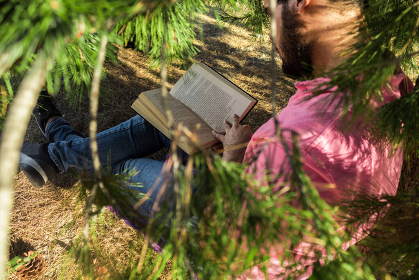 Kaukasischer Mann in Jeanshose und rosa Hemd sitzt an einem Baum im Wald und liest ein schwarzes Buch - Foto, Bild