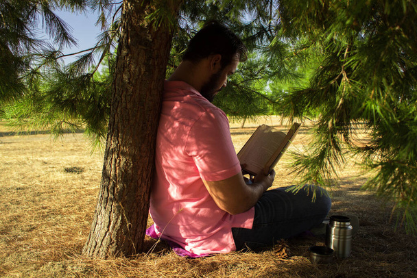 Caucasian man in jeans pants and a pink shirt sitting near a tree in a forest reading a black book - Photo, Image