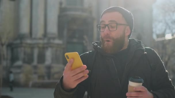 Funny excited man in glasses with paper coffee cup looking at phone screen while standing at street. Bearded guy using smartphone and rejoicing while having good news. Concept of win. - Video, Çekim