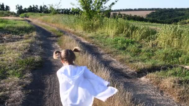 Meisje weglopen over landweggetjes midden in het zomers landschap. Langzame beweging. Achterzijde van een klein meisje dat 's zomers wegrent over een onverharde weg op het platteland. Witte cape vliegen in de wind. Blond paardenstaartje fladderend haar. Sereniteit van de kindertijd - Video