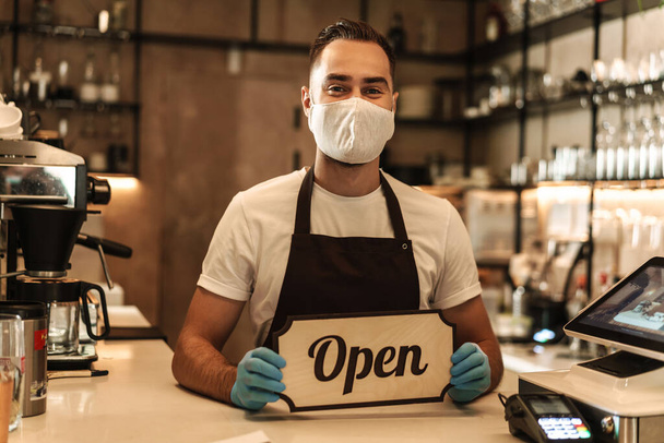 Man barista wearing medical mask showing open sign, standing behind coffee shop counter - Fotoğraf, Görsel
