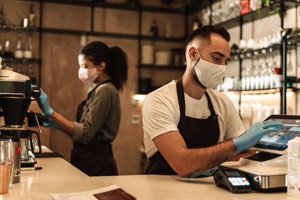 Two baristas wearing medical mask serving coffee behind the counter during quarantine indoors - Zdjęcie, obraz