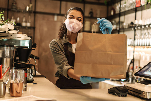 Happy woman barista wearing medical mask holding takeaway bag while standing behind the coffee shop counter - Foto, Bild