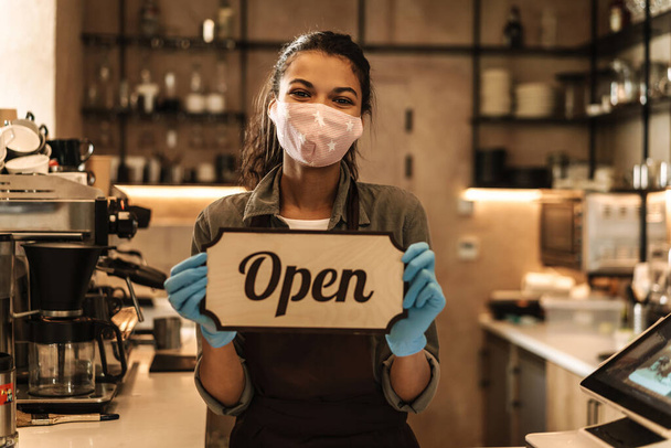 Happy woman barista wearing medical mask showing open sign while standing behind the coffee shop counter - Foto, Bild