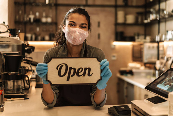 happy woman barista wearing medical mask showing open sign while standing behind the coffee shop counter - Foto, Imagem