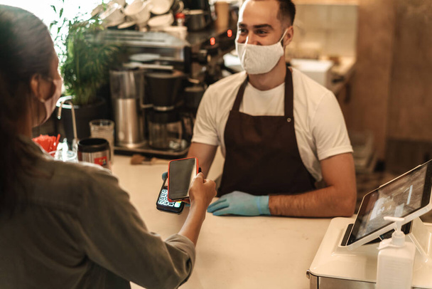 カウンターで注文を支払うためにキャッシュレス端末を保持する医療面マスクを身に着けている若い男バリスタ,携帯電話で支払う顧客 - 写真・画像