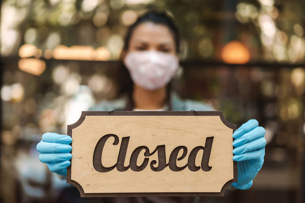 Woman wearing medical face mask holding closed sign while standing in front of a cafe - Foto, Imagen