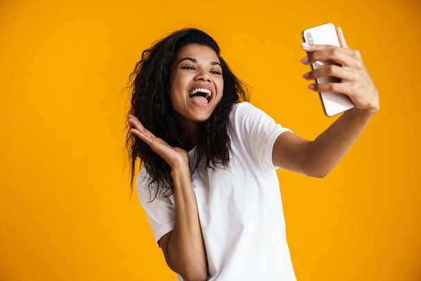 PSmiling young african woman taking selfie with mobile phone over yellow background - Valokuva, kuva