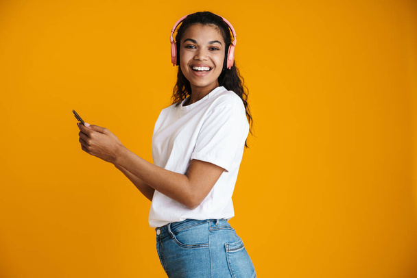 Imagen de una mujer afroamericana feliz con auriculares sonriendo y usando un teléfono móvil aislado sobre una pared amarilla - Foto, imagen