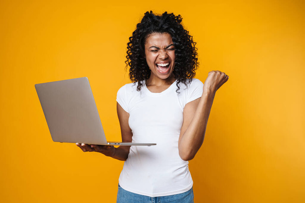 Bild der aufgeregten afrikanisch-amerikanischen Frau macht Siegergeste und benutzt Laptop isoliert über gelbem Hintergrund - Foto, Bild