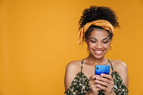 Képek boldog afro-amerikai nő mosolyog, és használja a mobiltelefon izolált sárga fal - Fotó, kép